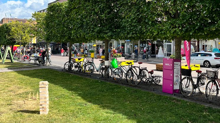 Pifflbox bredvid cyklar och spel i parkmiljö, med människor och butiker i bakgrunden mitt i Linköpings sommartorg. Piffl