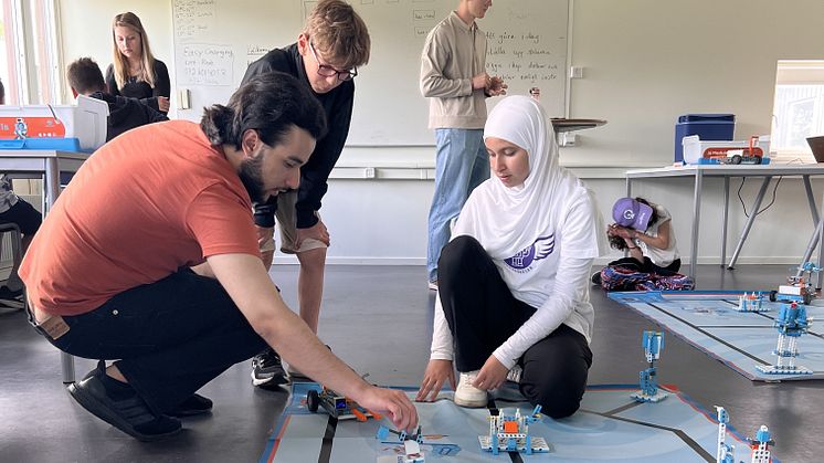 En grupp människor deltar i en robotskola där de bygger och programmerar robotar. En person i orange tröja och en i vit hijab