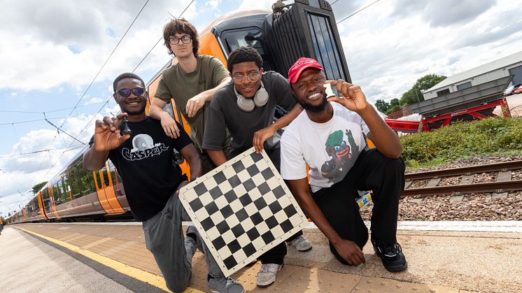 Four men posing in front of a West Midlands Railway train with chess pieces and a chess board