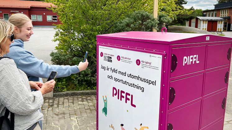Två personer står vid en rosa Pifflbox utomhus och använder sina mobiltelefoner för att låna aktiviteter i Uppsala.