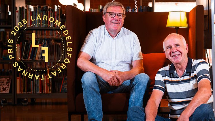 två män sitter bredvid varandra, den ena i en fåtölj, den andra på golvet. De ler och tittar in i kameran.