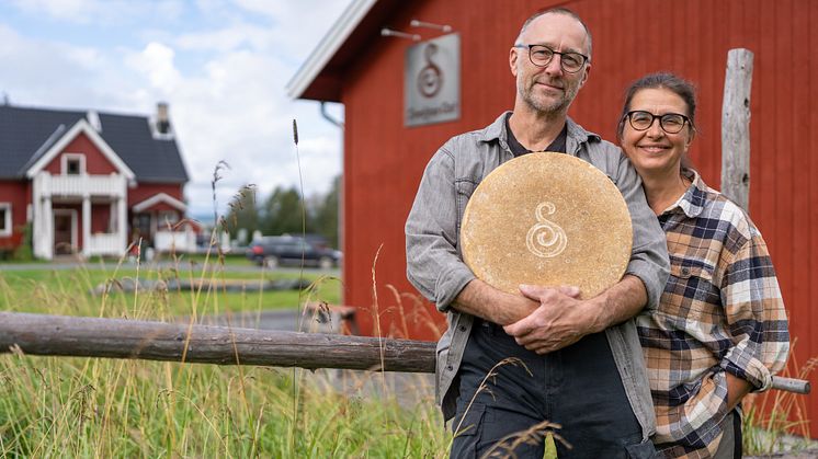 En man och en kvinna framför ett rött hus. Mannen håller i en ost.