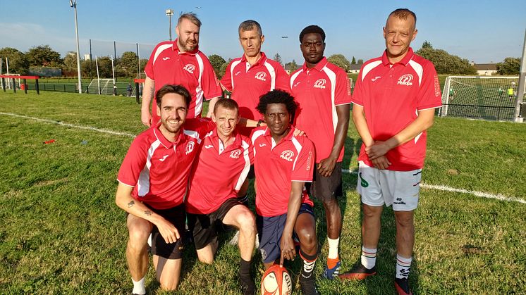 Photo shows a sunny playing field with a team of rugby players wearing red and white rugby shirts
