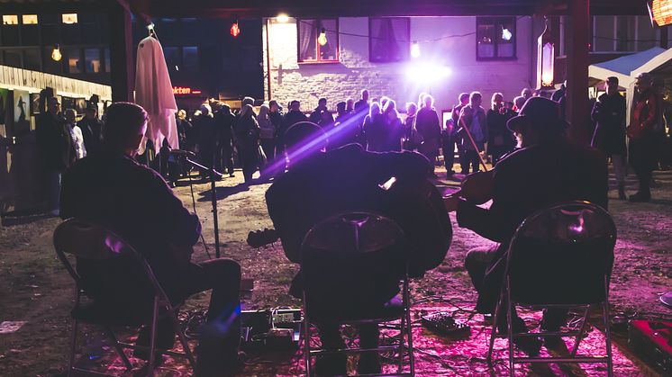 Tre musiker sitter med ryggarna mot kameran och publik framför på en bakgård, det är kväll och lila ljus i bilden