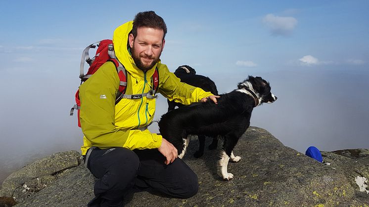 Bearded man wearing yellow jacket with his dog on top of a mountain