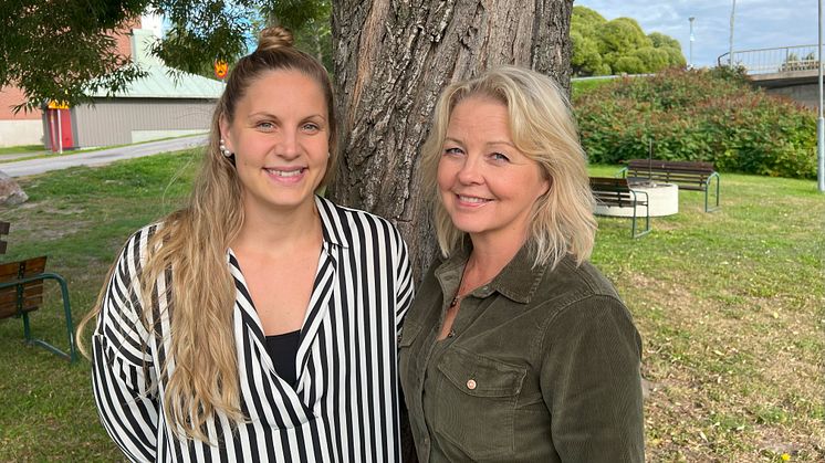 Matilda och Camilla framför ett träd. Grönt i bakgrunden och del av bro. Båda ler.