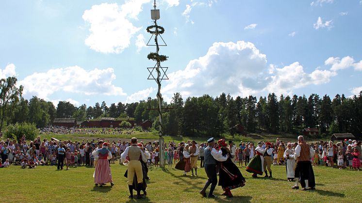 Dansande par klädda i folkdräkt som dansar runt midsommarstången på ängen vid Vallby Friluftsmuseum. Många människor ser på.