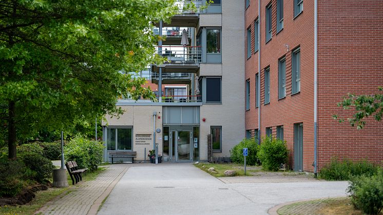 Entré till ett äldreboende med flera våningar, framför huset finns bänkar och stora träd.