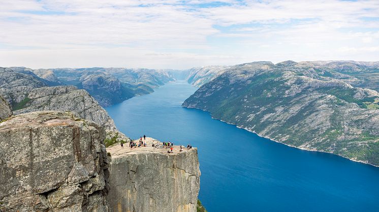 A mountain overhang with people on top. The long fjord 600 meters down from plateau. Mountains on each side of fjord.