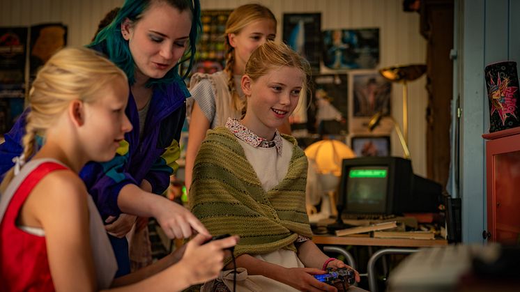 En eldre ungdom utkledd i 90-tallsklær viser tre mindre jenter hvordan de spiller på et gammelt dataspill. Foto: Anno museum