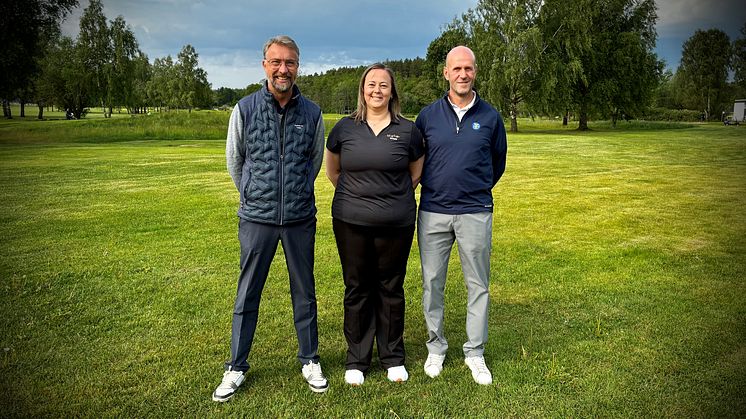 Tre personer stor på en grön gräsmatta. De ser glada ut och håller händerna bakom ryggen.