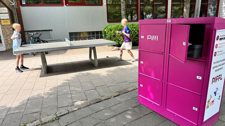 Två barn spelar pingis vid ett pingisbord intill en rosa Piffl aktivitetsbox. Boxens skåpdörrar är öppna, redo för utlåning.