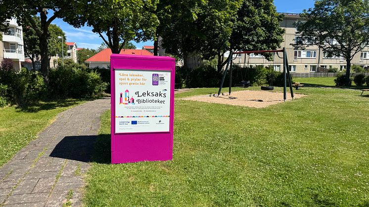 En rosa Pifflbox från Leksaksbiblioteket står vid en gångväg i en grön park med gungställning i bakgrunden.