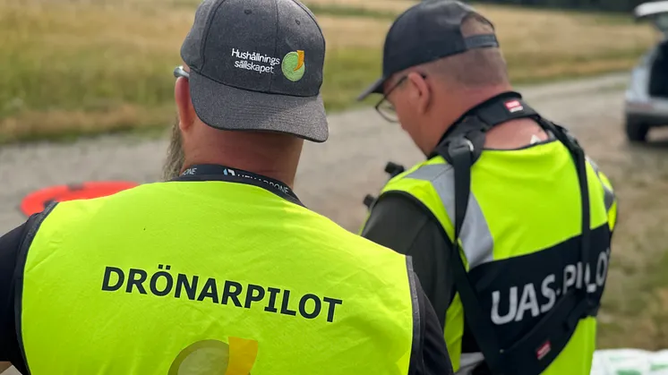 Två drönarpiloter i varselvästar vid ett jordbruksfält. På västarna står det 'DRÖNARPILOT' och 'UAS.PILOT'.