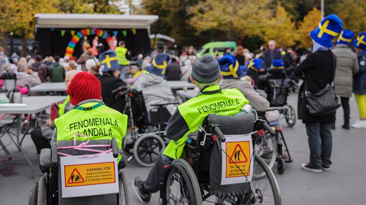 I förgrunden två personer i rullstol som tittar mot en scen med en regnbågsfärgad båge. 