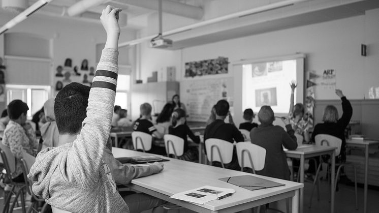 Elever sittter i ett klassrum, några av dem räcker upp handen