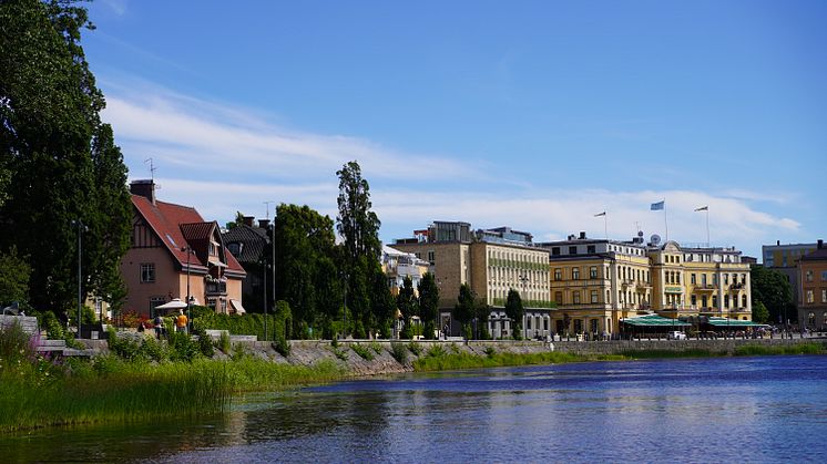Klarälven i förgrunden och stadshotellet i bakgrunden en solig sommardag med frodig grönska, blå himmel, små lätta moln. 