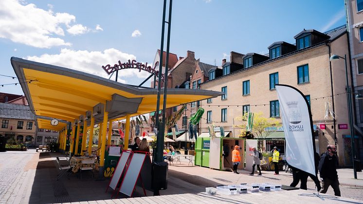 Gula busstak och evenemangsflaggor på torget kring Botulfsplatsen, människor som går och pratar. 
