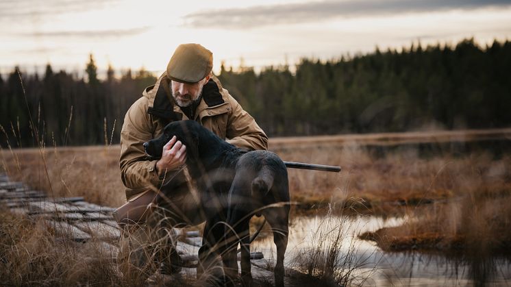 Man i jaktkläder från Pinewood klappar en svart hund vid en spång i höstligt myrlandskap.