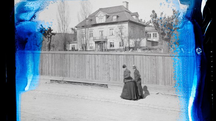 Svartvitt foto på två damer i sekelskifteskläder som promenerar på en gata framför ett hus. Bilden har blåaktiga skador.