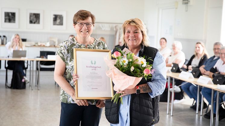 Två personer med ett inramat diplom och en blombukett. I bakgrunden syns flera människor som sitter.