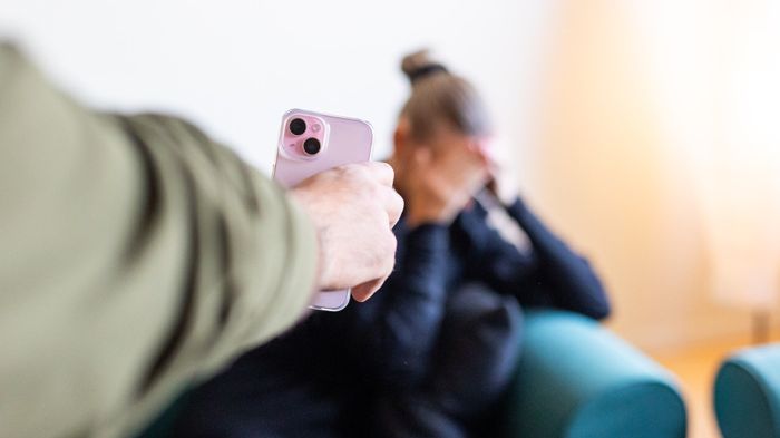 En man håller upp en mobil och visar den för en kvinna som håller för sitt ansikte. Kvinnan sitter ned och mannen står upp. Man ser mannens arm och hand. 