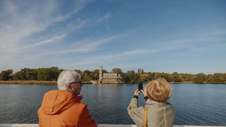 Eine Dame und ein Herr fahren mit dem Schiff die Havel entlang. Sie fotografieren die Heilandskirche Sacrow.