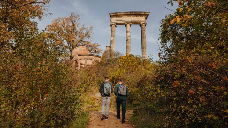 Zwei Männer gehen einen sandigen Weg entlang. Dieser führt zwischen herbstlichen Gewächsen durch. Ruinenberg im Hintergrund.