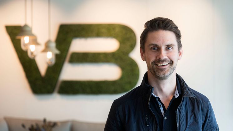 Erling Pettersen smiler foran en vegg med VB Gruppens logo i grønt. Han har mørkt hår, kort skjegg og er kledd i mørk jakke o