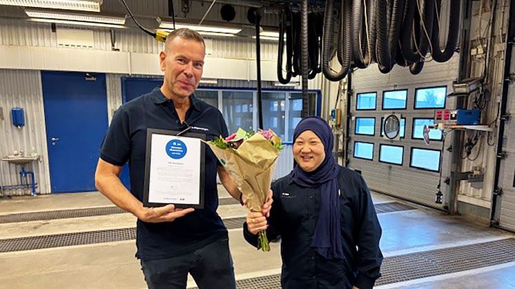 En man håller ett diplom och tar emot en bukett blommor av en kvinna i en besiktningshall.