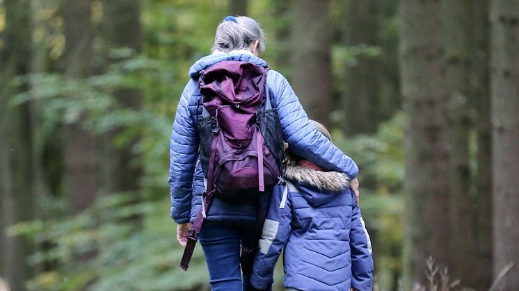 En kvinna går i skogen och håller armen om ett barn. De syns bakifrån. 