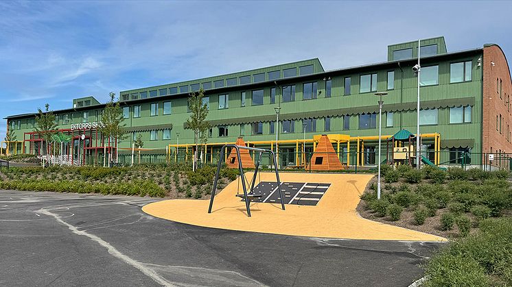 Fastighet med grön fasad med texten Ektorps skola över entrén. Kompisgunga och gröna buskar i förgrunden.