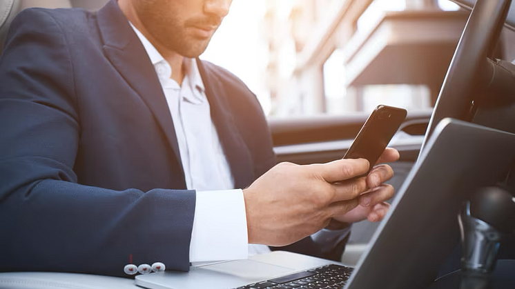 A man in a suit, sitting in a car, with a laptop and a smartphone