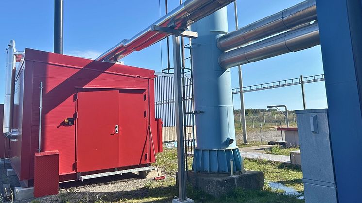 En röd sjöfartscontainer som innehåller en ny bioljepanna. Flera stålgrå rör och en stor, blå fjärrvärmeskorsten syns också.