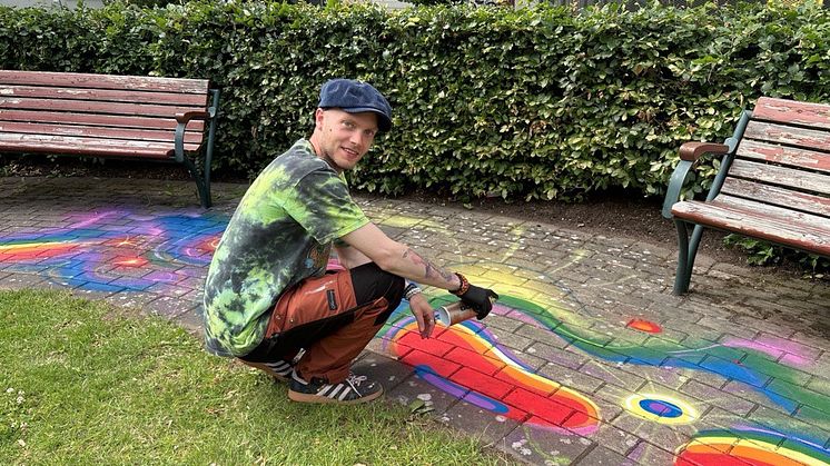 Man med keps tittar in i bilden och håller i en sprayburk, han har målat marken med regnbågsfärger 