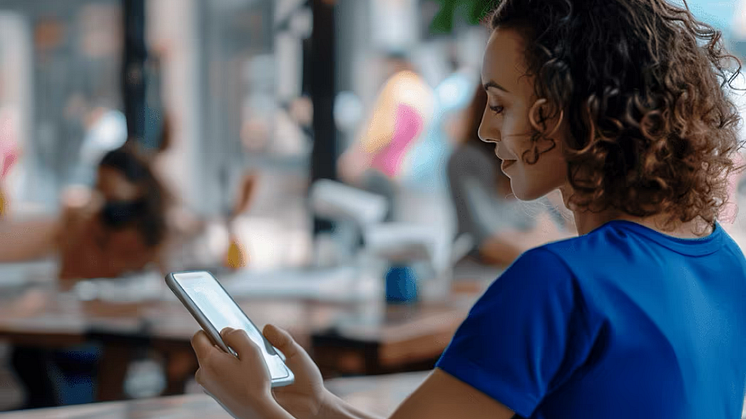 A woman in a blue shirt, with curly dark brown hair, sitting at a table in a restaurant, looking at a smartphone in her hands