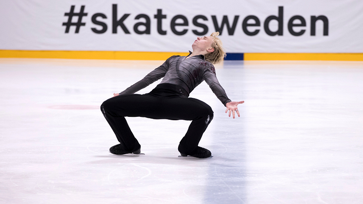 Konståkare i bakåtlutad pose på isen, med utsträckta armar och #skatesweden i bakgrunden