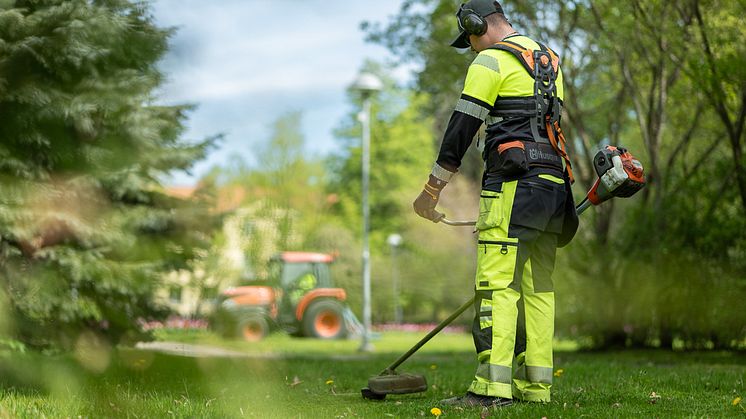 En man i gula arbetslkäder står i en park med en grästrimmer. I bakgrunden syns en röd traktor. Parkarbete verkar pågå.
