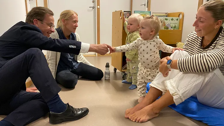 En man i kostym sitter på golvet tillsammans med två små barn och deras mammor. 