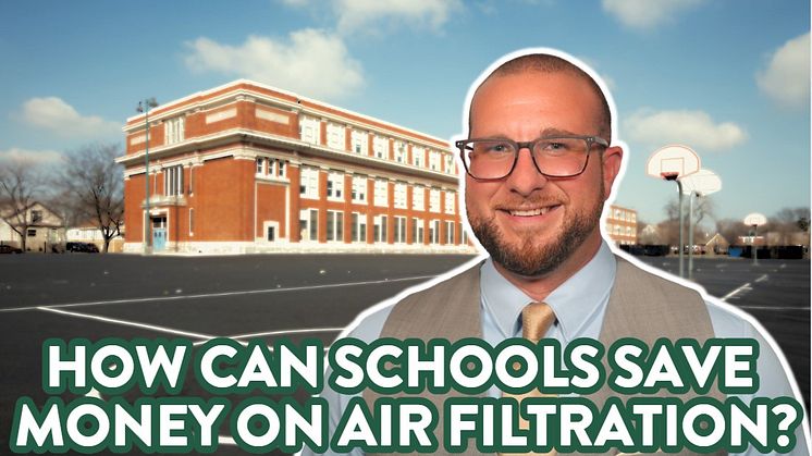 A man with glasses stands in front of a school. It partially cloudy. A blue sky is visible. 