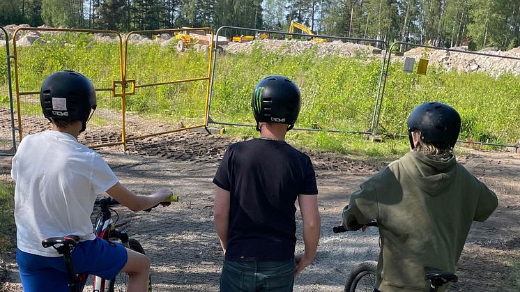 Tre barn i cykelhjälmar blickar ut över byggarbetsplats.