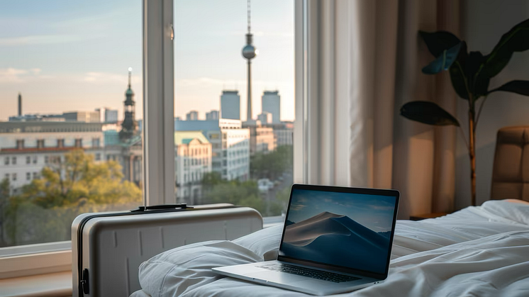 Inside of a hotel room, a laptop on the bed, a suitcase next to it. Outside the window a cityscape 
