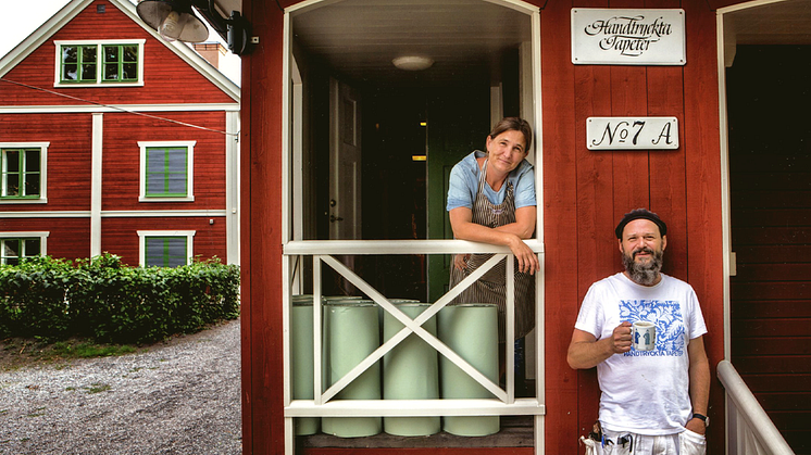 Two people at the entrance of a red building marked "Handtryckta Tapeter".