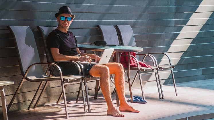 En man i shorts sitter vid poolen med laptop i knät och jobbar. 