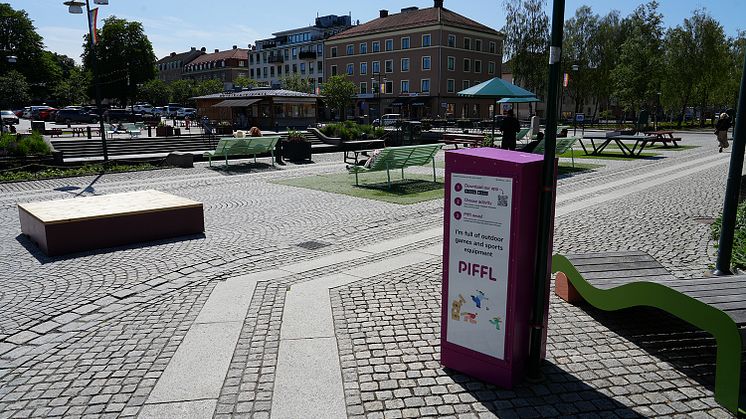 En rosa Pifflbox står i en stadspark med kullersten i Alingså. Runt omkring syns gröna bänkar, picknickbord, växter och männi