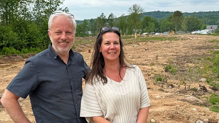 Två personer framför ett byggnadsområde där det grävs i marken. I bakgrunden syns skog och byggnader.