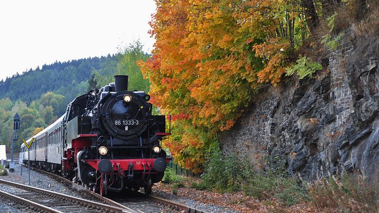 zu sehen ist eine alte Dampflok, die auf Schienen durch die bunte Natur des Herbstes fährt