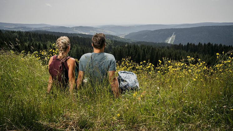 Ein Mann und eine Frau sitzen auf einer blühenden Wiese und schauen in die Ferne der Mittelgebirgslandschaft.