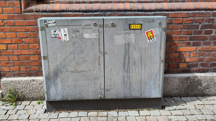 Elskåp i metall med klistermärken, placerat på kullersten intill en tegelfasad.