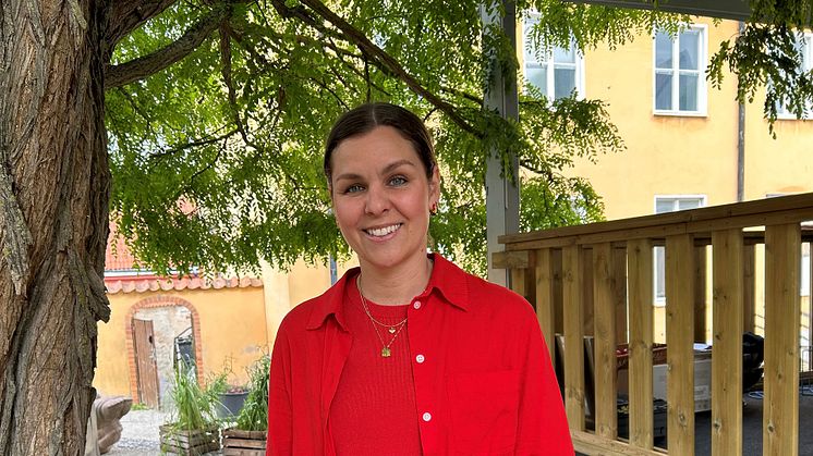 Lina Ejlertsson står under ett lövträd och ser in i kameran. Hon har på sig en röd tröja, röd skjorta och vita byxor.